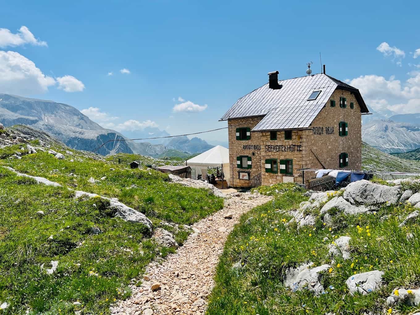 Rifugio Bien Seekofelhütte stone mountain hut on grassy trail with rocky peaks under blue sky.