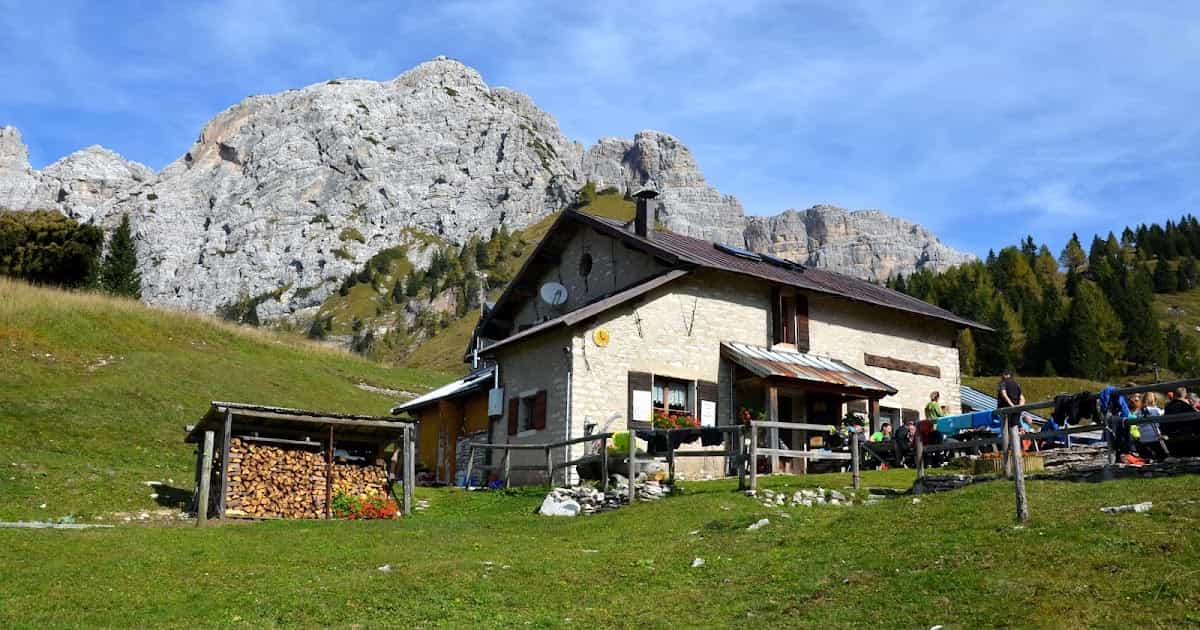Rifugio Boz Dve mountain hut with hikers resting below large rocky peaks and a woodpile.