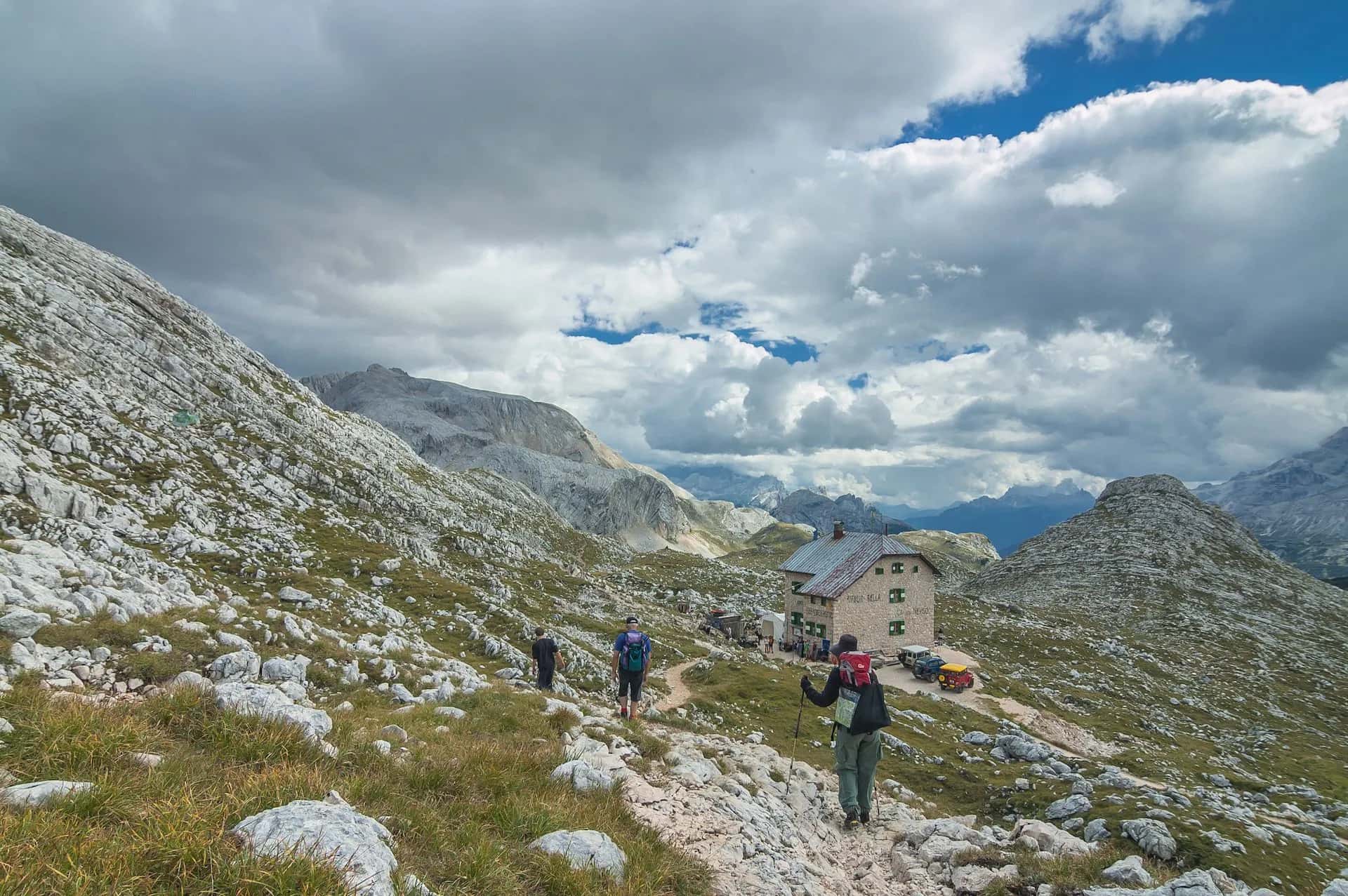 Hikers approaching Rifugio Biella mountain hut among rocky terrain under a dramatic cloudy sky.