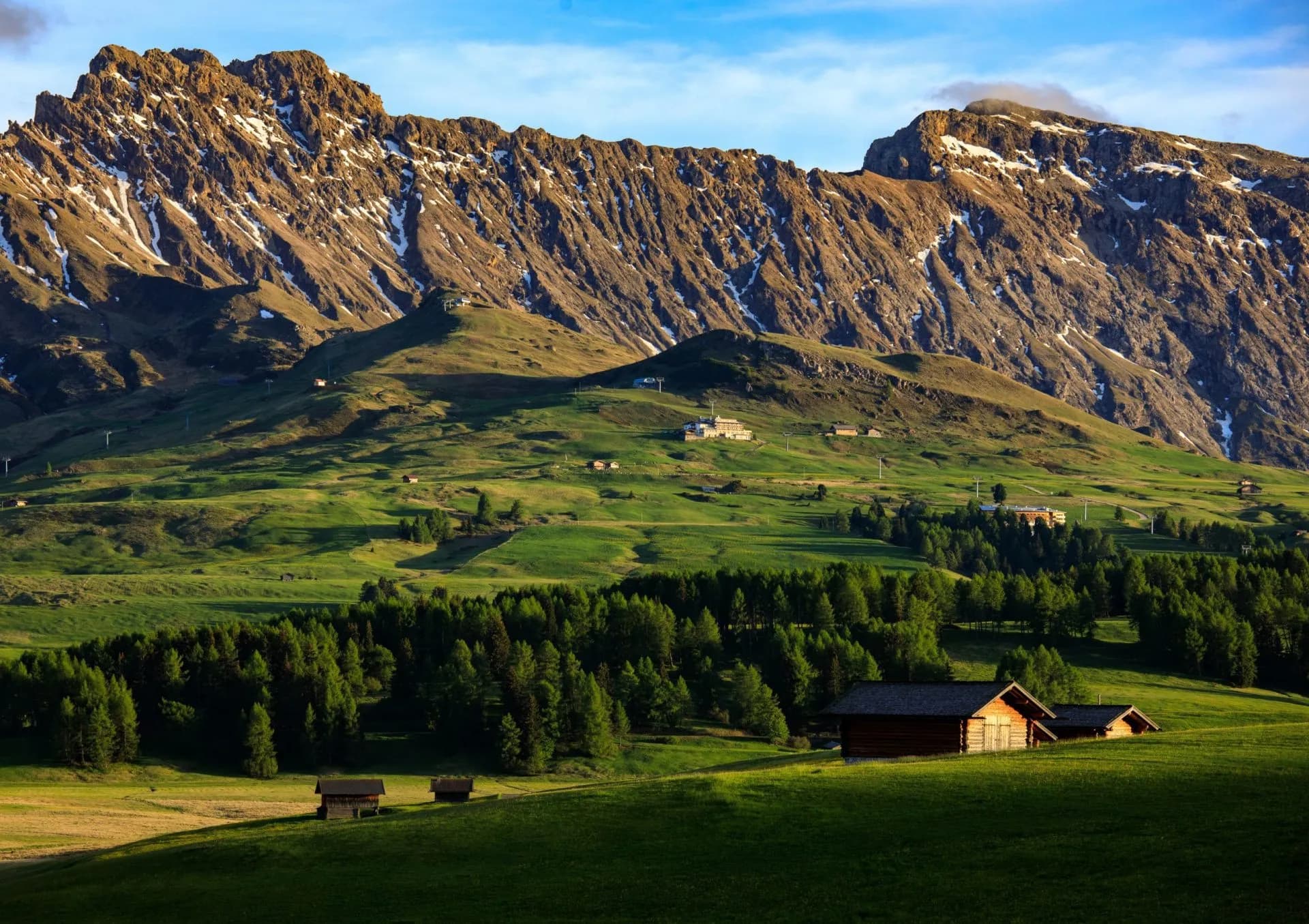 Alpe di Siusi rolling green meadows with wooden huts below rugged, partially snow-dusted mountains.