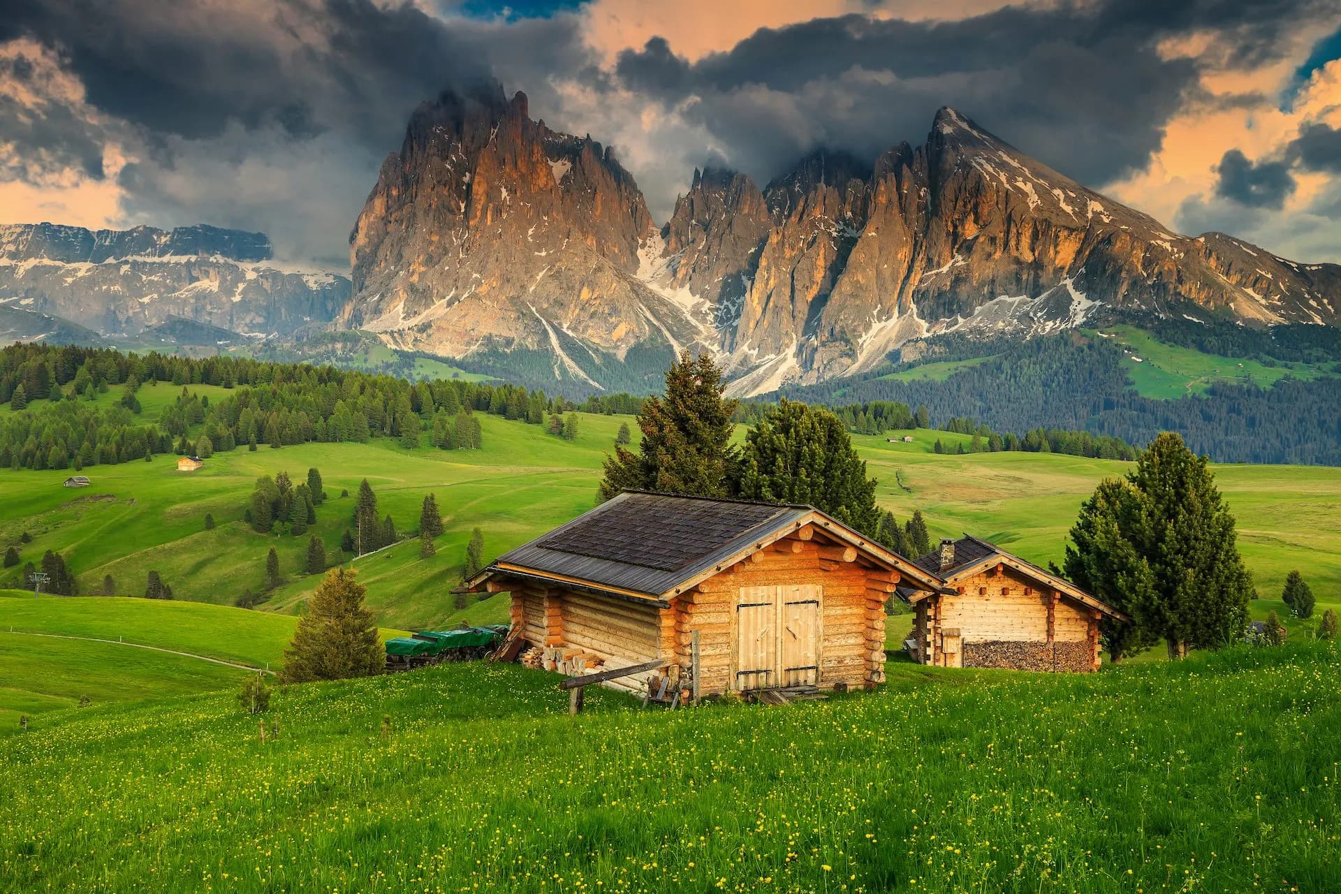 Wooden alpine huts in green meadow below dramatic mountains under cloudy sky, Seiser Alm.