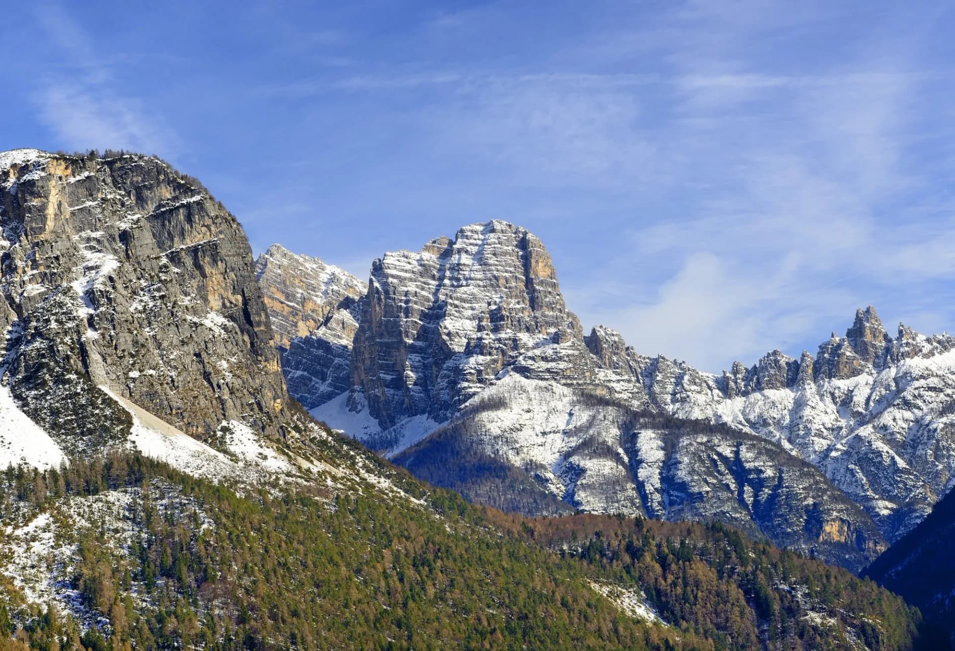 Snow-capped mountains rising above forested slopes under a blue sky in Forno di Zoldo.