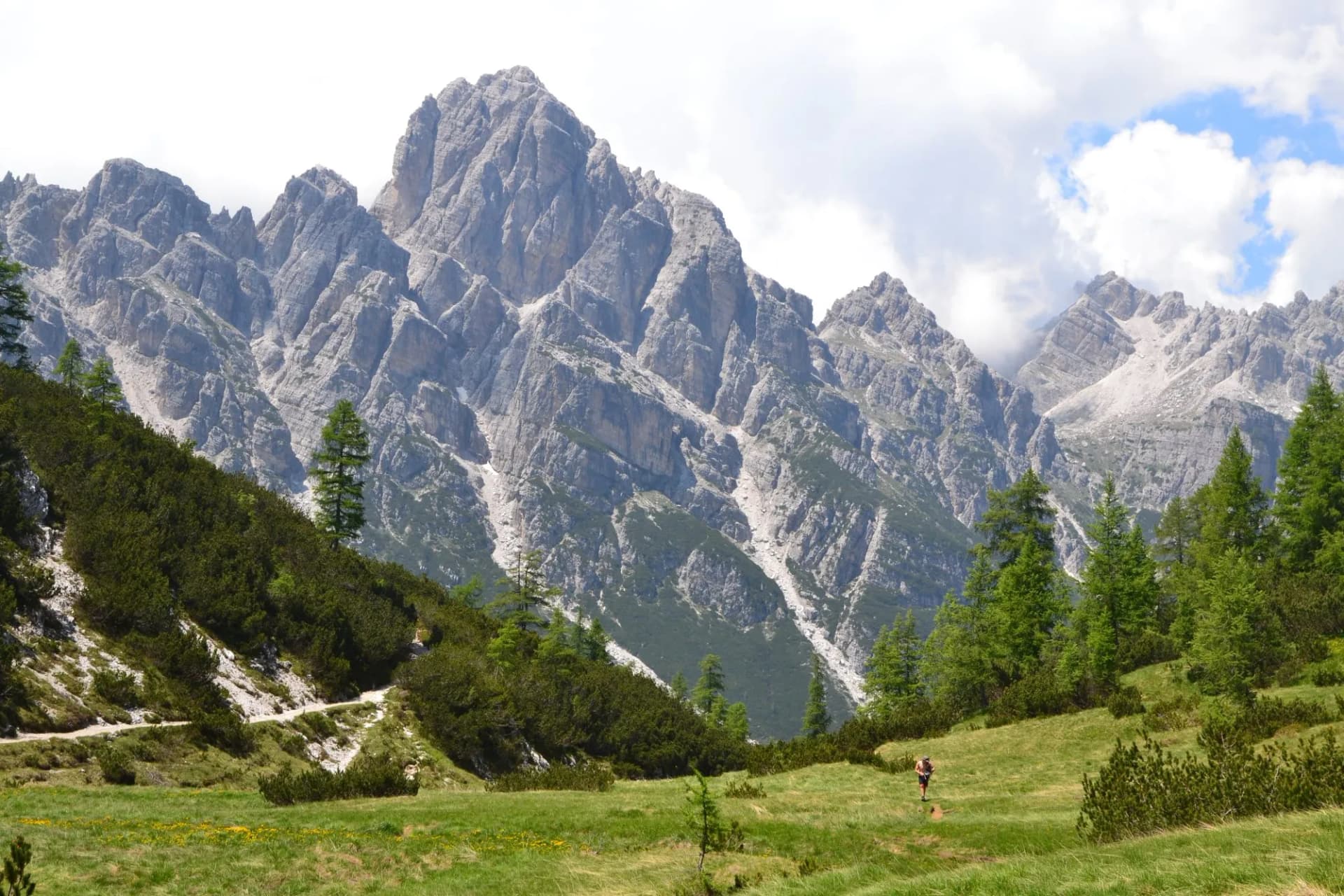 Hiker on grassy trail below jagged limestone peaks in Dolomites above Forno di Zoldo.