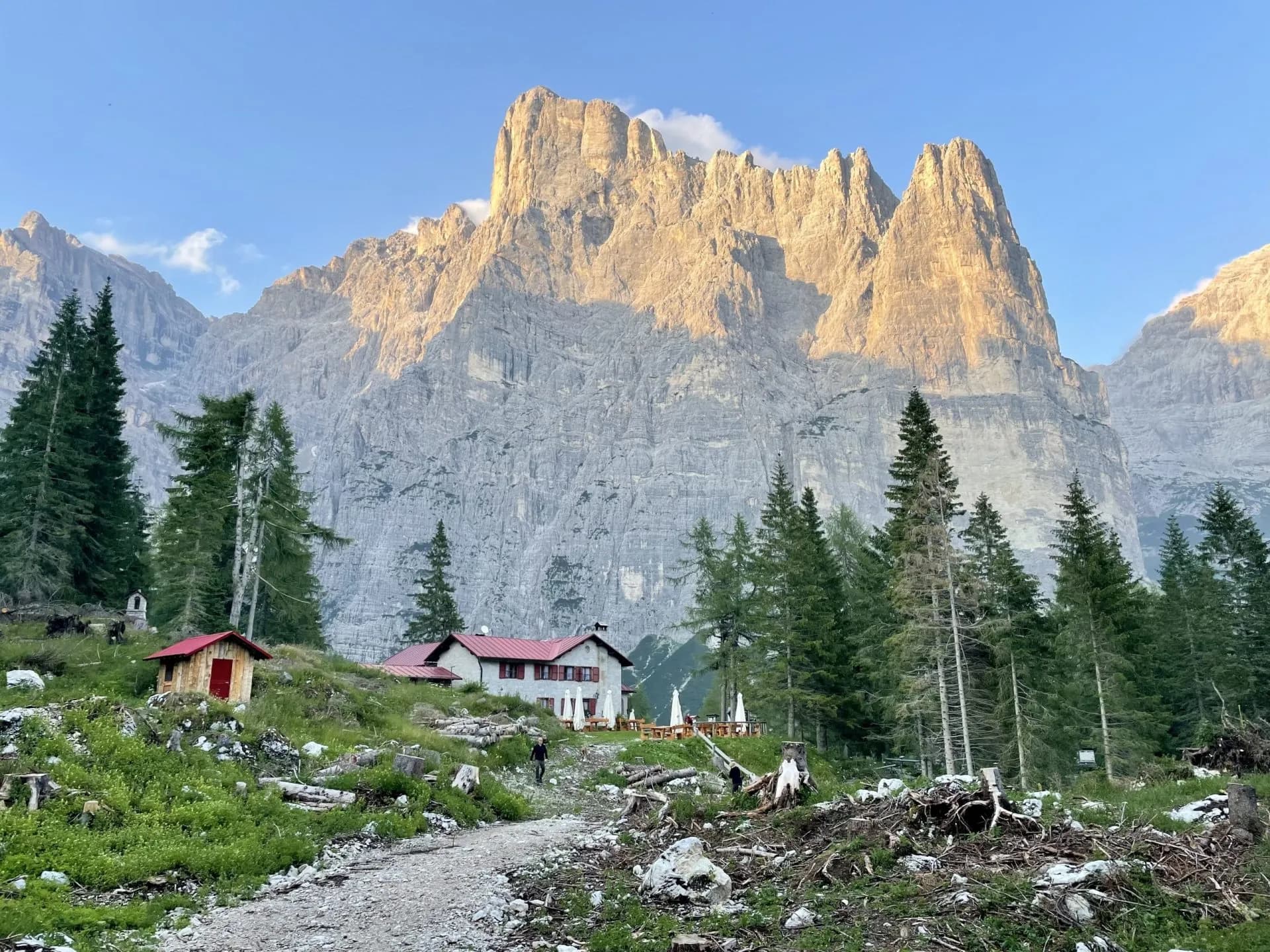 Mountain hut with red roof below massive sunlit rock face and pine trees