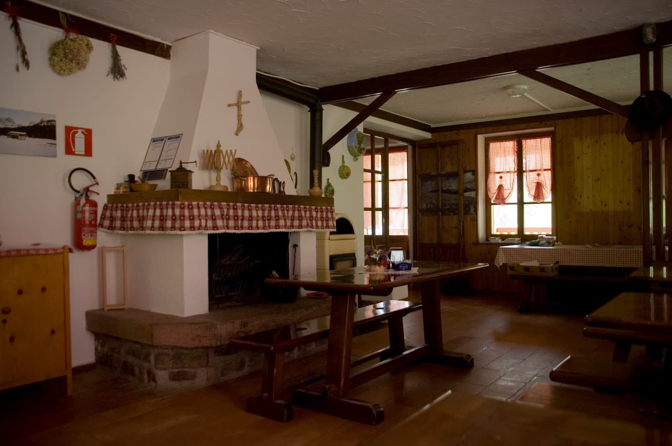 Rustic dining room interior with large stone fireplace, wooden tables, and checkered curtains at Rifugio Furio Bianchet.