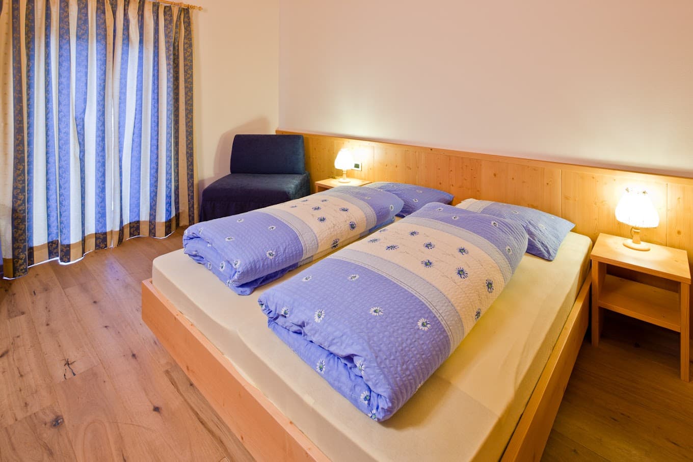 Bedroom in Berggasthaus Pederü with wooden frame bed, blue floral duvets, and striped curtains.