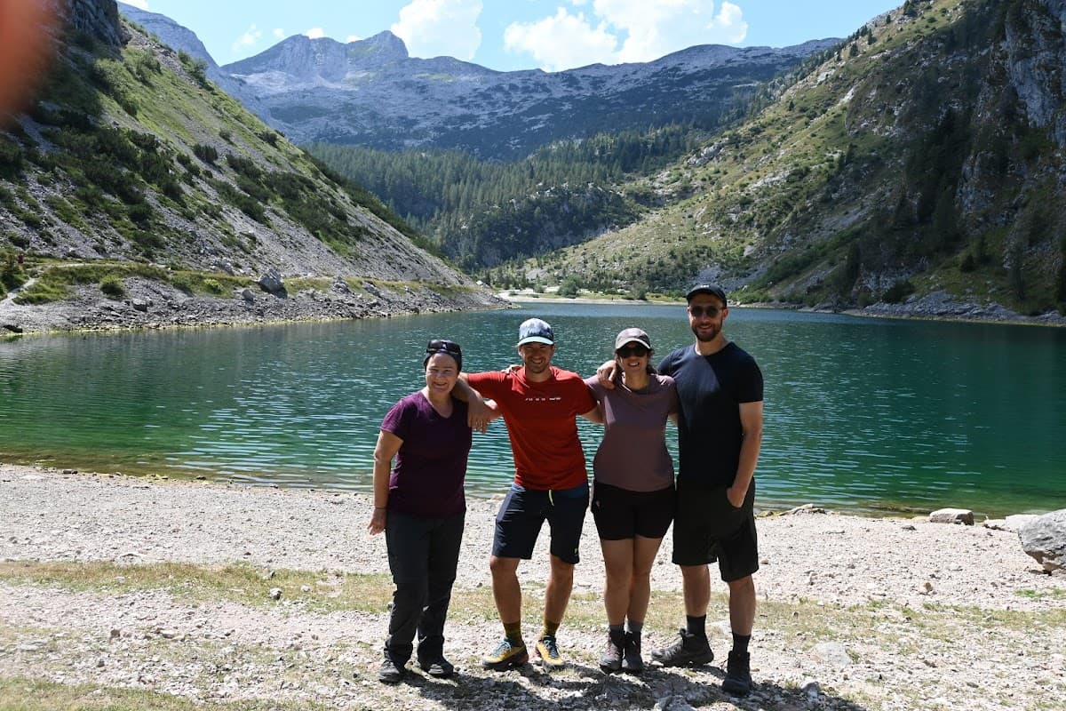 Four hikers posing by a clear green alpine lake surrounded by rocky, forested mountains