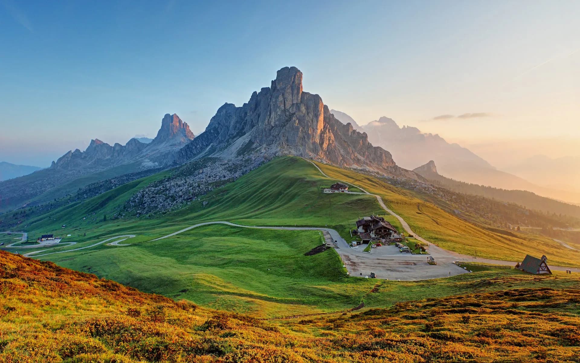Passo Giau mountain pass with jagged peaks, green slopes, and buildings at sunrise or sunset.