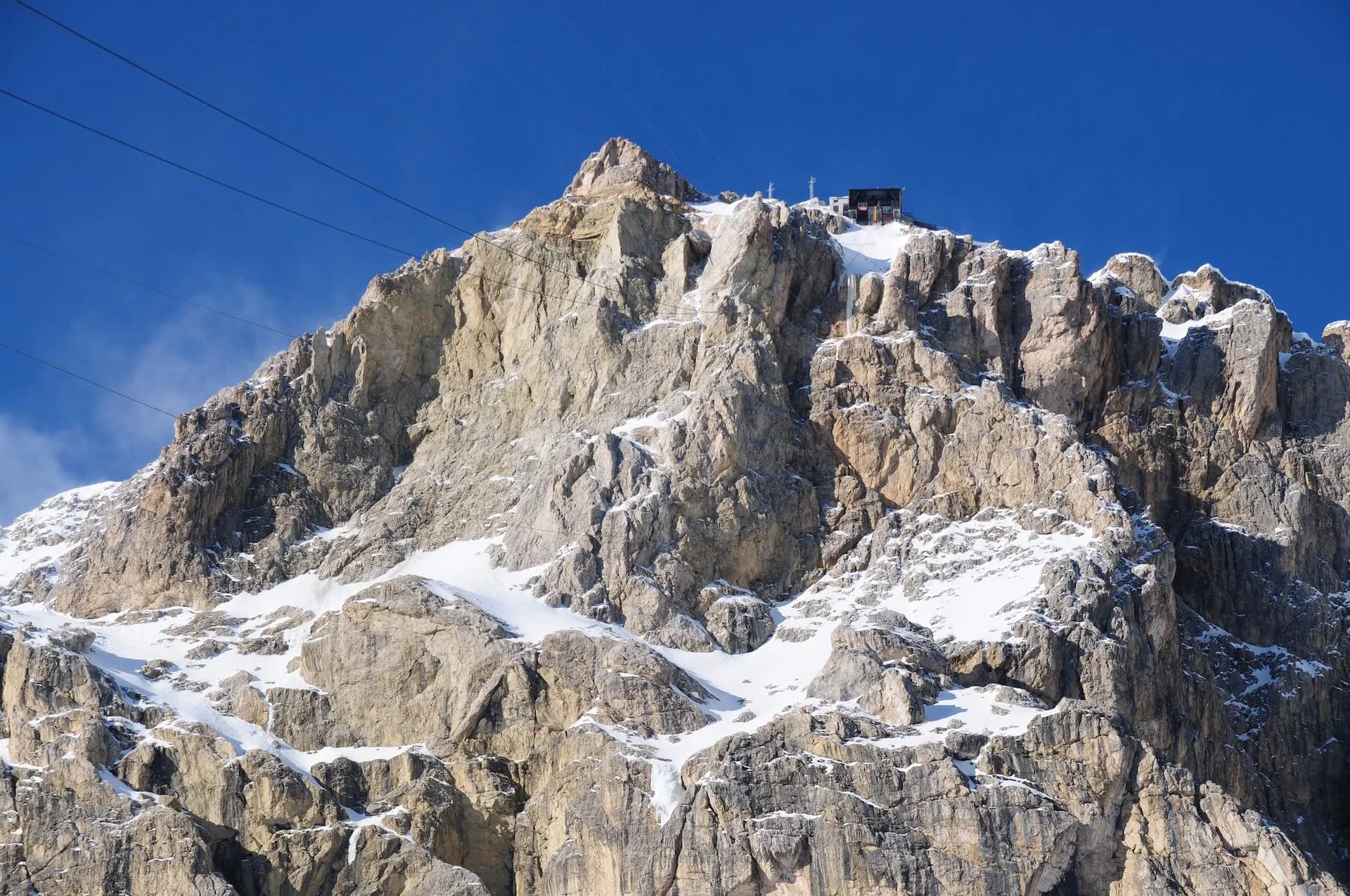 Rocky mountain peak with snow patches, a summit building, and cable car wires against a blue sky.