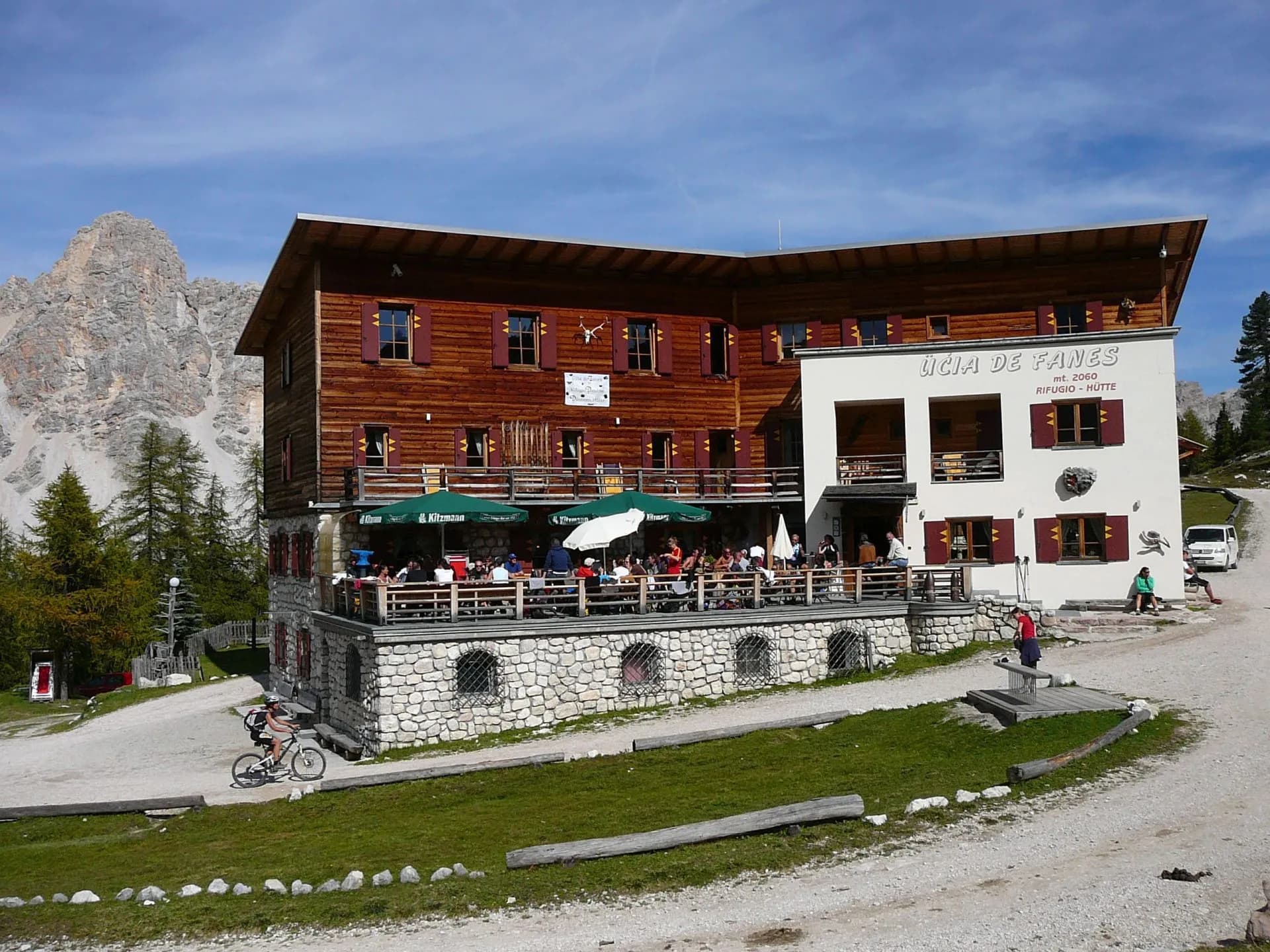 Mountain hut with outdoor dining, cyclist arriving, rocky peak in background at Fanes.
