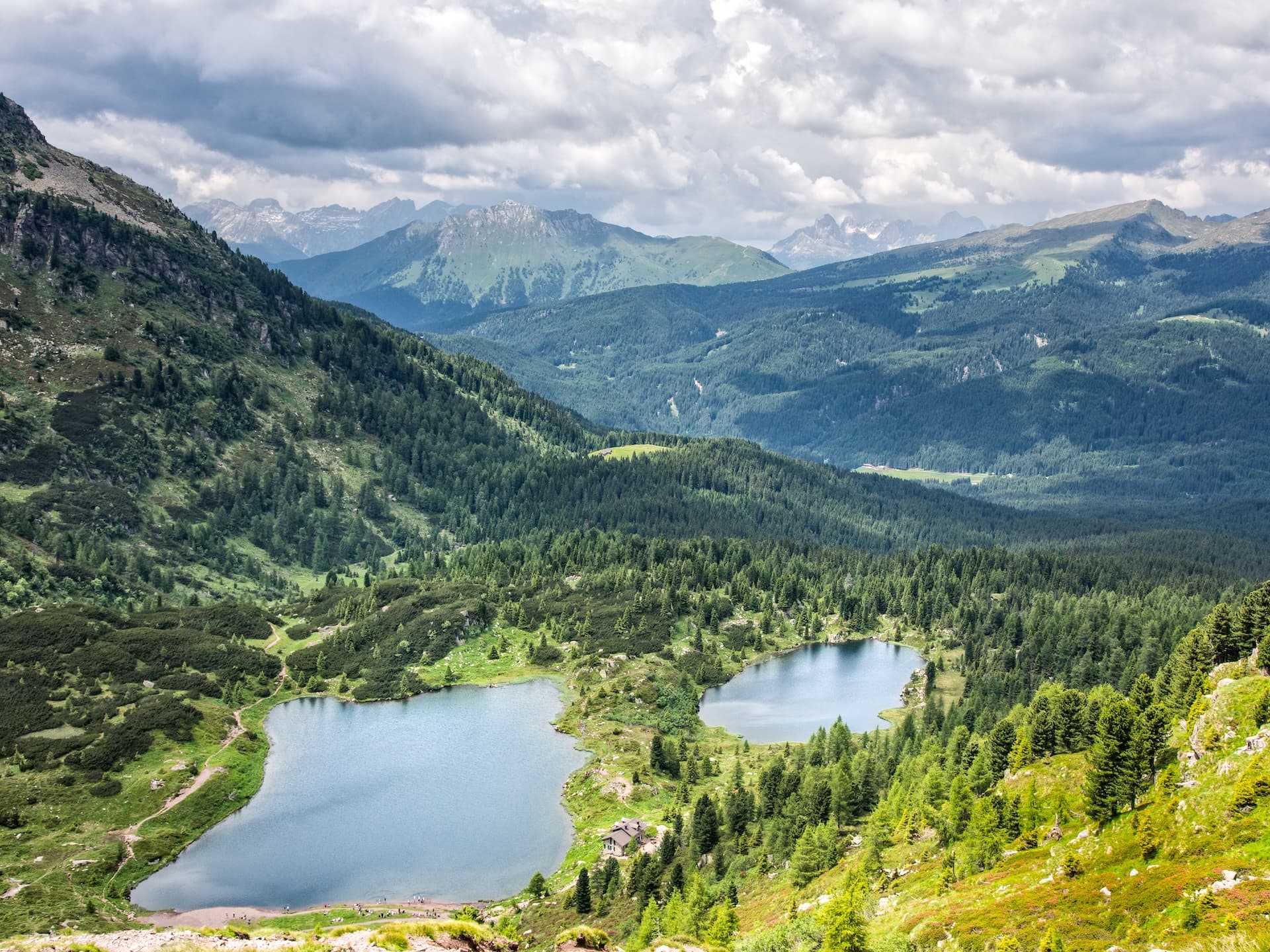 Two alpine lakes nestled in a valley surrounded by dense pine forests and distant mountains under a cloudy sky.