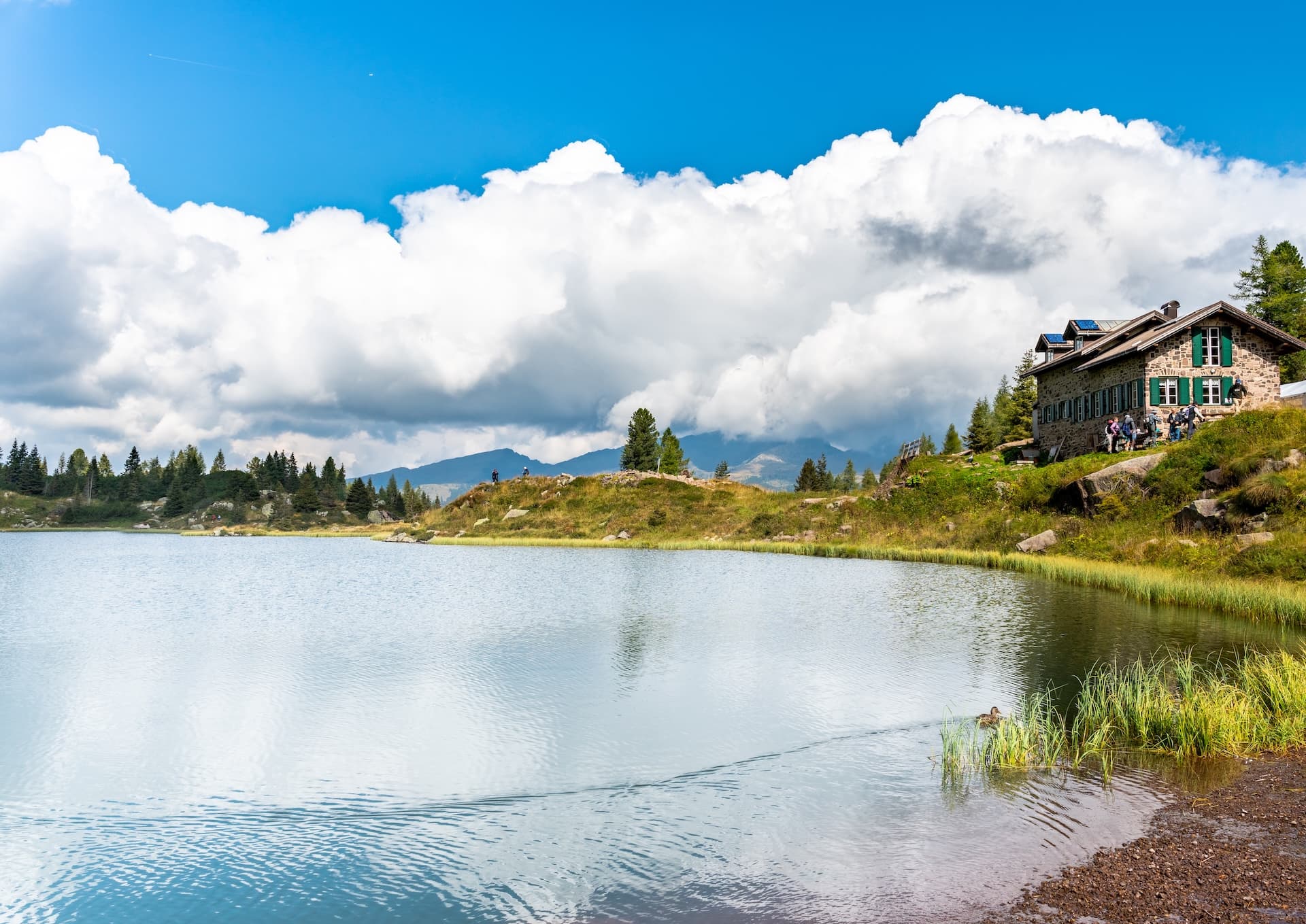 Alpine lake with stone Rifugio and hikers, mountains under dramatic clouds