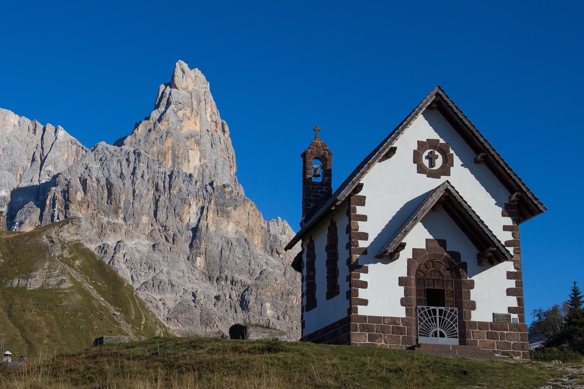 Small white and stone chapel in alpine meadow beneath towering rocky mountain peaks.