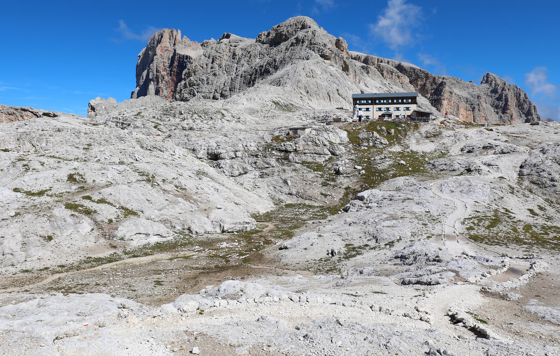 Rifugio Rosetta mountain hut nestled in rocky terrain below large Dolomite peaks under blue sky.