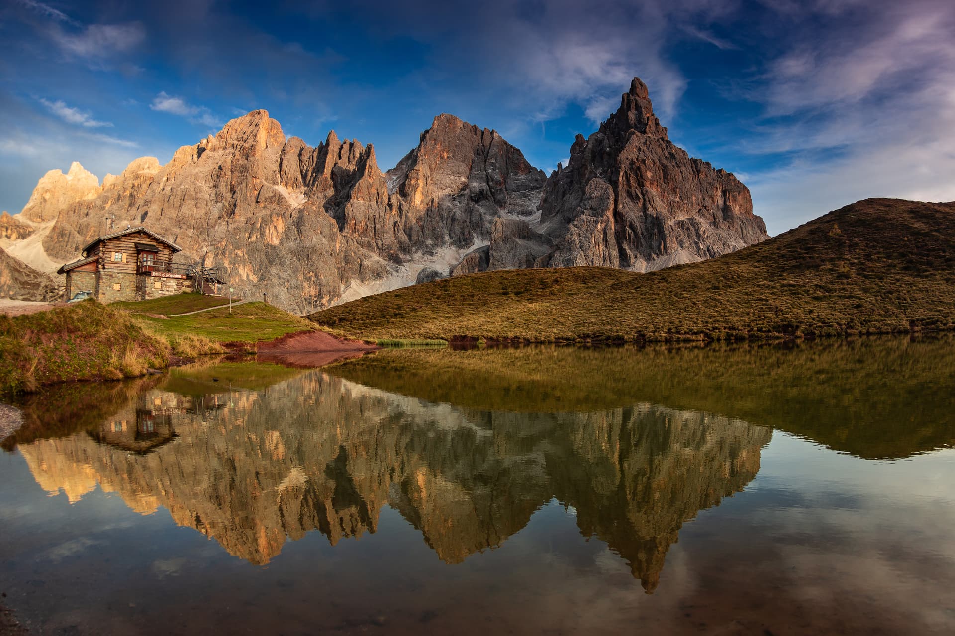 Alpine lake reflecting jagged mountain peaks and a wooden refuge hut under a blue sky at Passo Rolle.