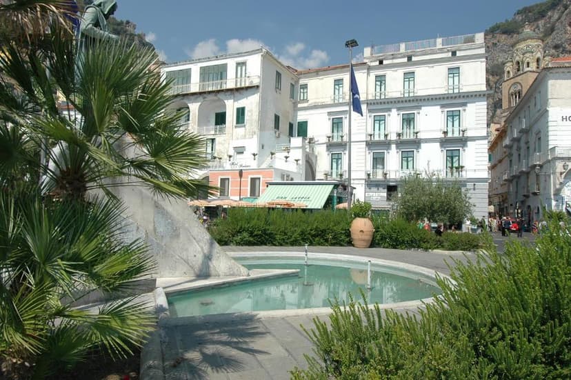 Fountain with palm trees and white buildings in a sunny coastal town square.