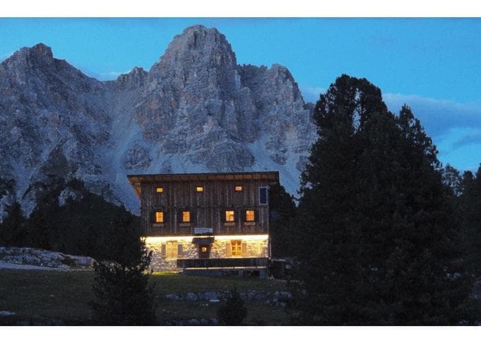 Illuminated wooden mountain hut at dusk below large rocky peaks in the Dolomites.