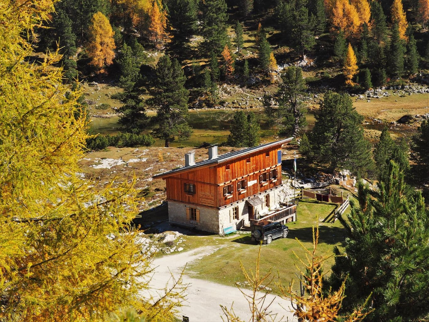 Wooden mountain hut with stone base near a small pond, surrounded by autumn trees.