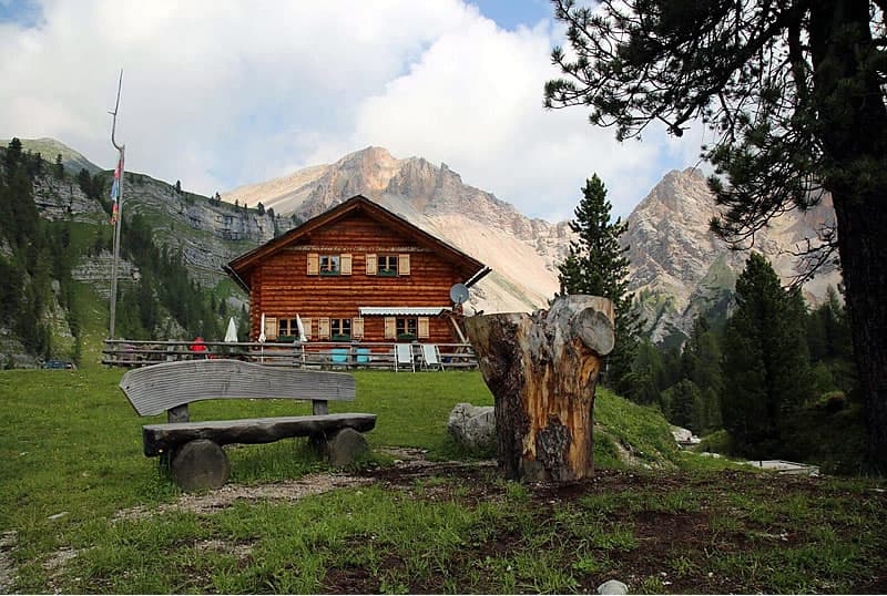 Wooden mountain hut with log bench in front of steep, rocky peaks.