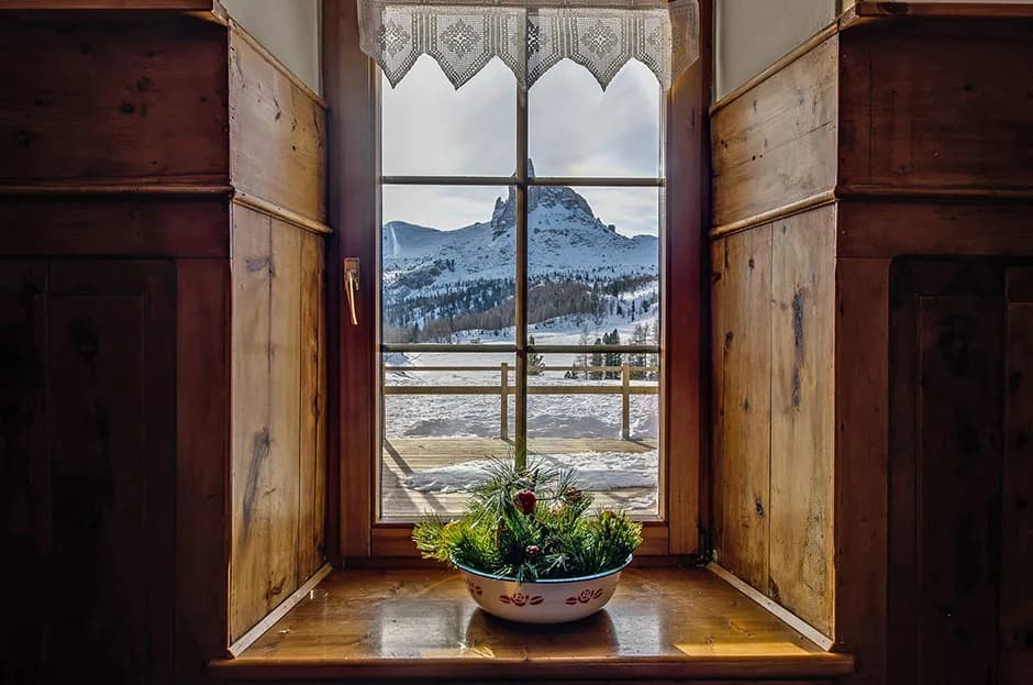 Window view of Rifugio Croda da Lago with snow-covered mountains and pine centerpiece.