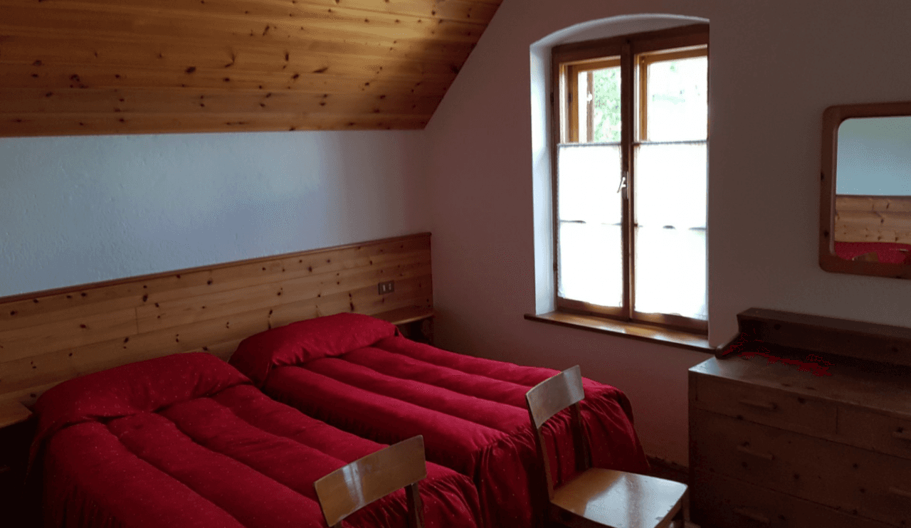 Twin bedroom with red bedding, wood paneling, and bright window in Rifugio 5 Torri.