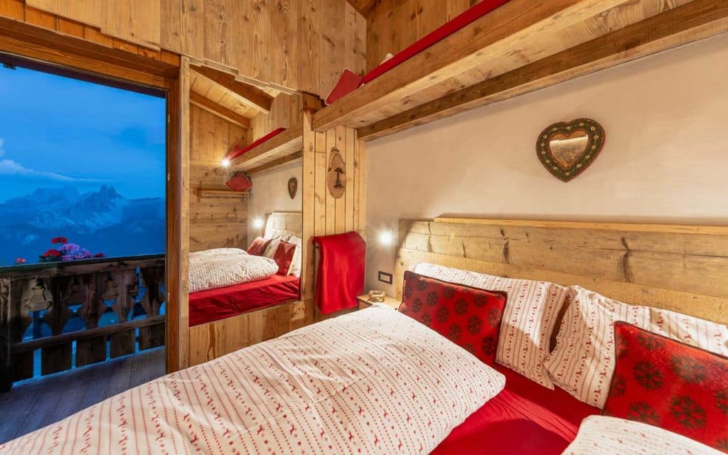 Cozy wooden bedroom with red bedding and balcony view of blue mountains at Rifugio Pomedes.