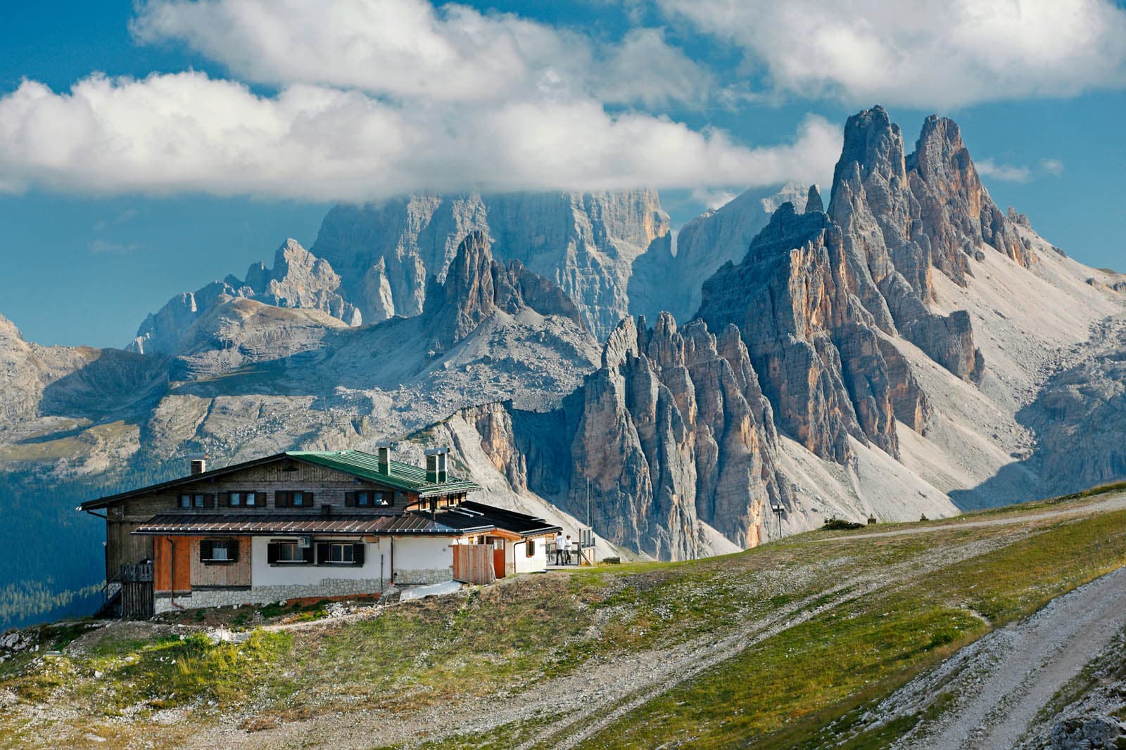 Rifugio Pomedes mountain hut below jagged, towering peaks in the Dolomites under a cloudy blue sky.