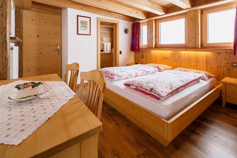Cozy wooden hotel room with double bed, dining area, and bright windows at Berghotel Passo Giau.