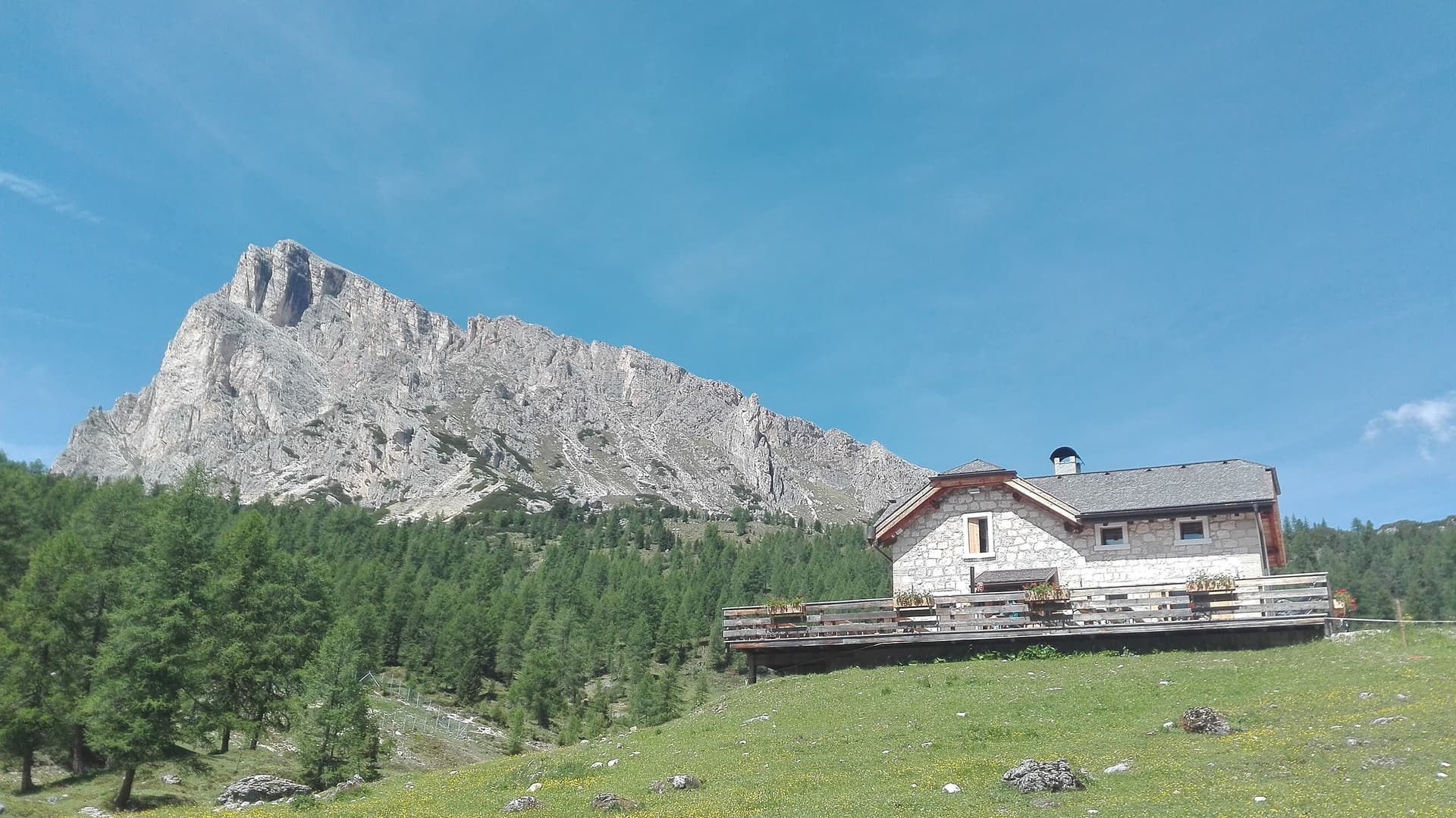 Stone mountain hut with wooden deck below a rocky peak and pine forest, Malga Giau.