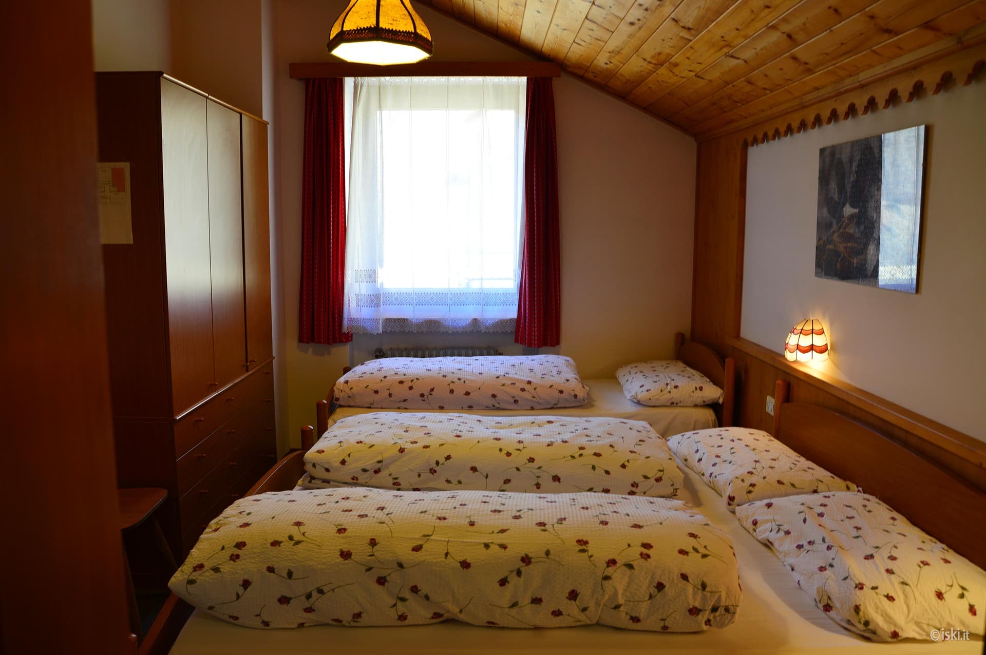 Bedroom with multiple beds, wooden ceiling, and bright window with red curtains at Rifugio Fedare.
