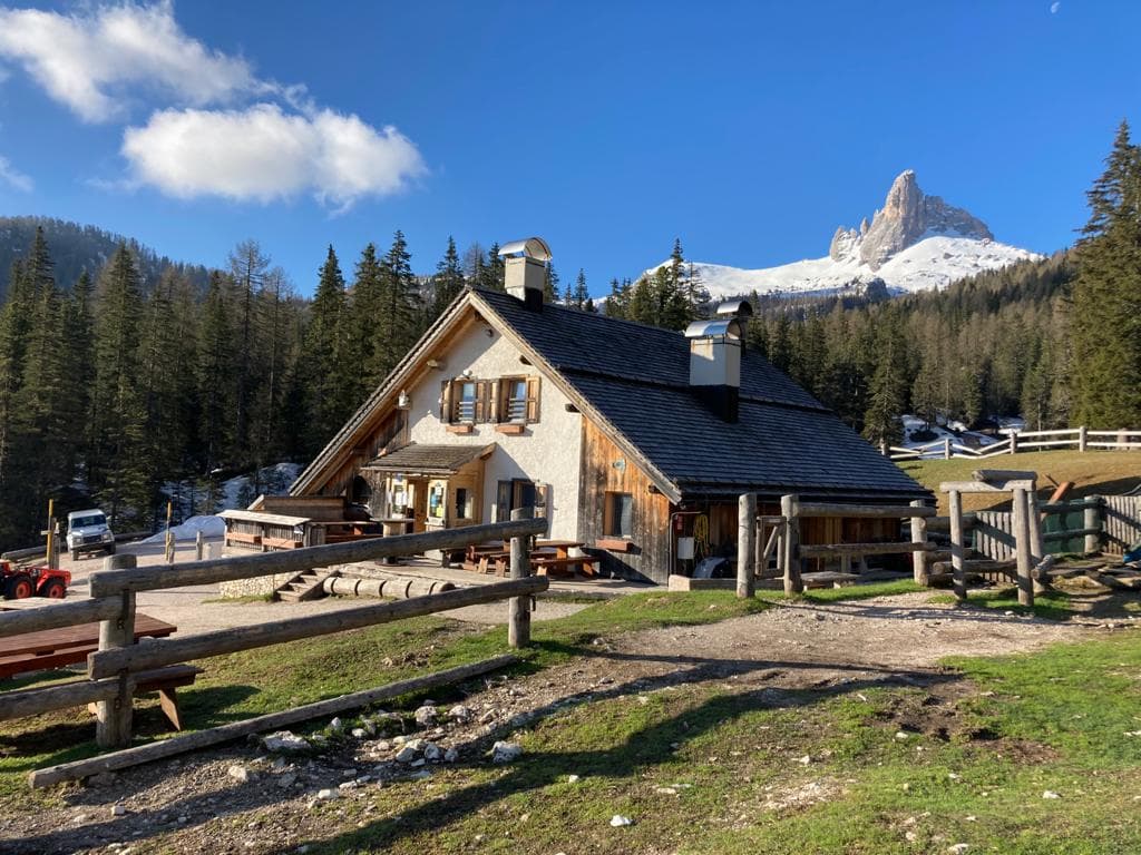 Mountain hut Malga Federa with snow-capped Dolomite peak in background under blue sky.