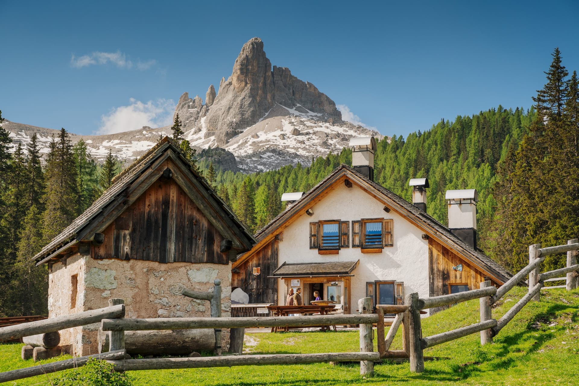 Alpine hut with wooden fence in front of rocky, snow-dusted mountain and green forest