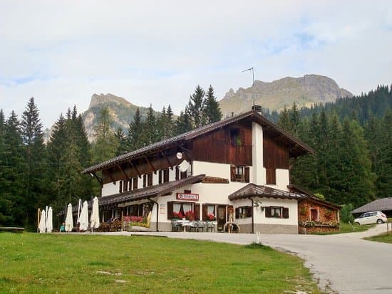 Rifugio Aquileia building with outdoor seating, surrounded by pine forest and mountains.