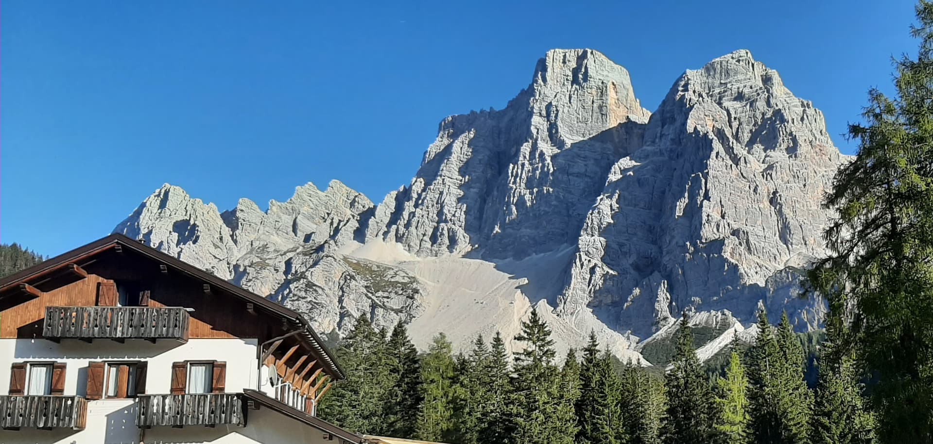 Alpine lodge with wooden balconies beneath rugged, sunlit mountains and clear blue sky.
