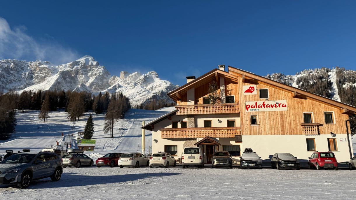 Rifugio Palafavera building in snow with parked cars and snowy mountains.