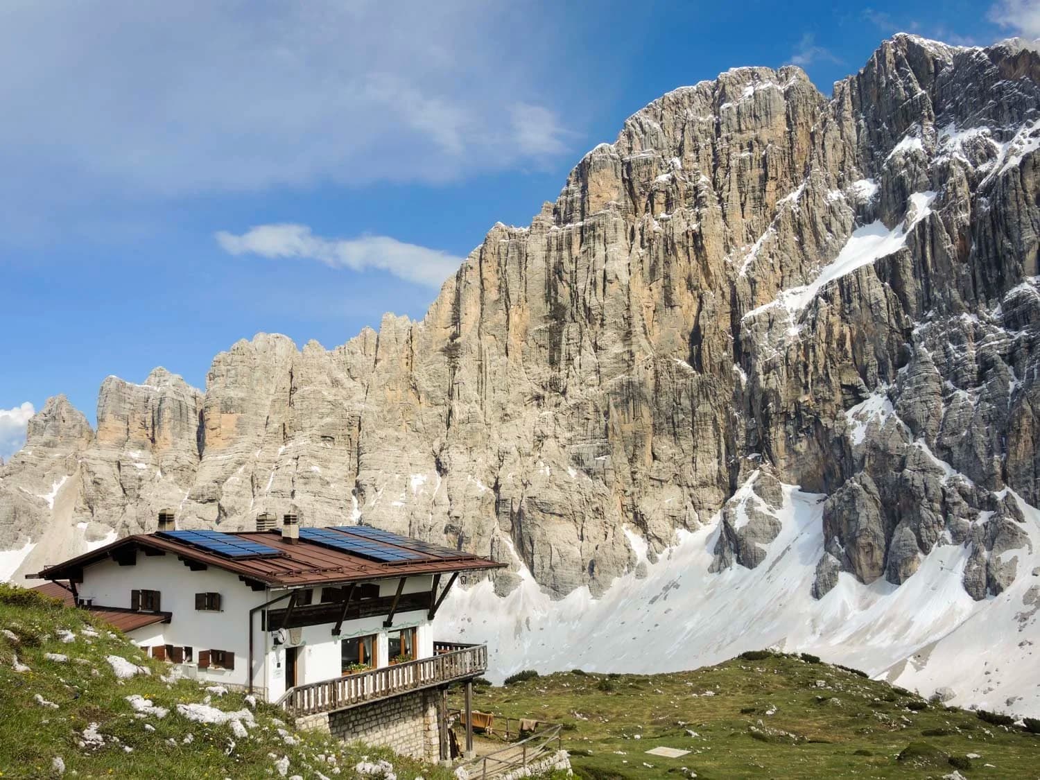 Rifugio Tissi mountain hut below sheer rock face with residual snow under blue sky