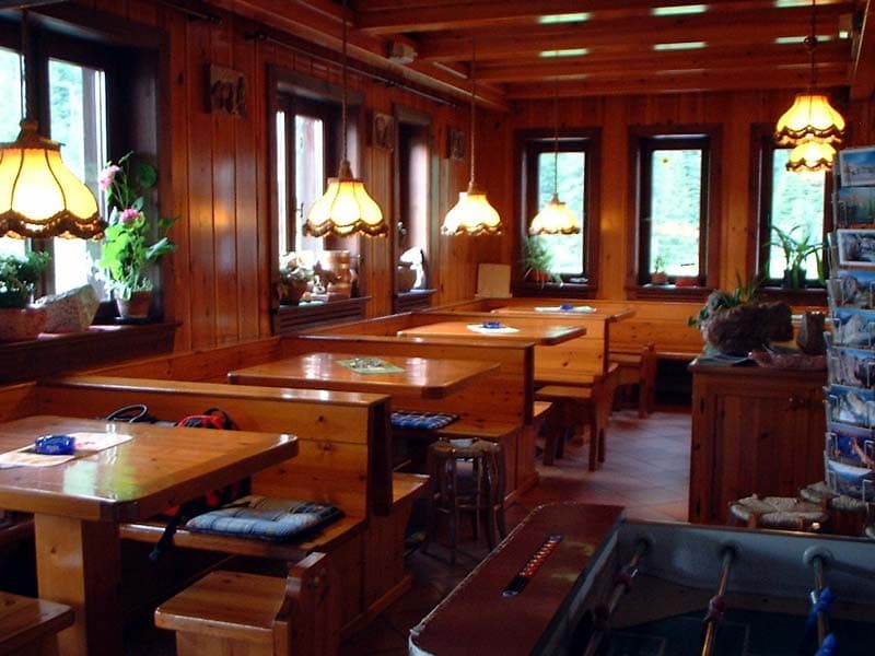 Rustic wooden interior dining area with booths, hanging lamps, and windows showing greenery. Rifugio San Sebastian.
