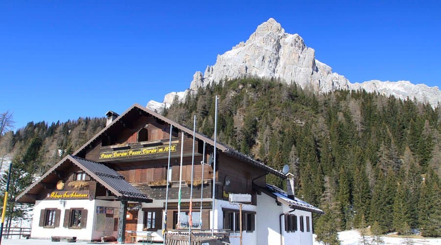 Rifugio San Sebastiano lodge below rocky mountain peaks and pine forest in winter.