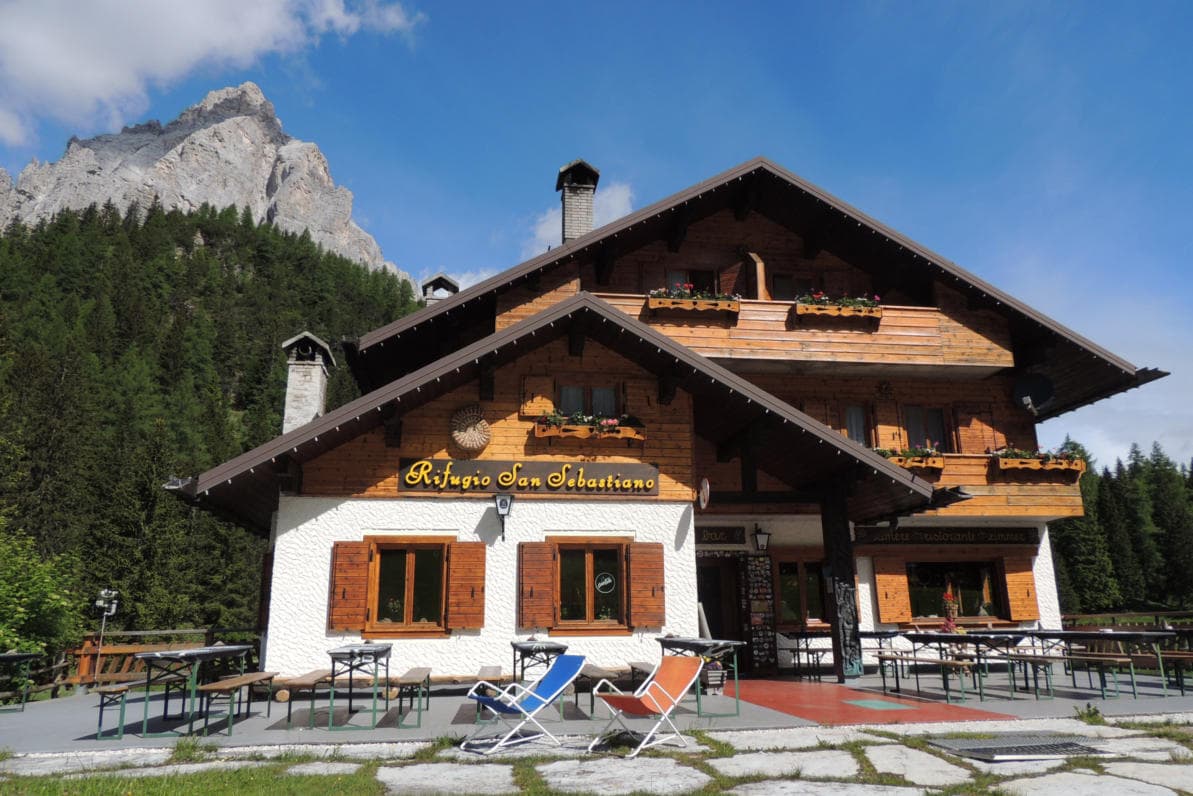 Rifugio San Sebastiano mountain lodge with outdoor seating against a backdrop of a rocky peak and forest.