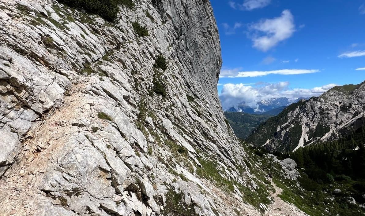 Narrow hiking trail with safety cable along a steep limestone rock face in the mountains.