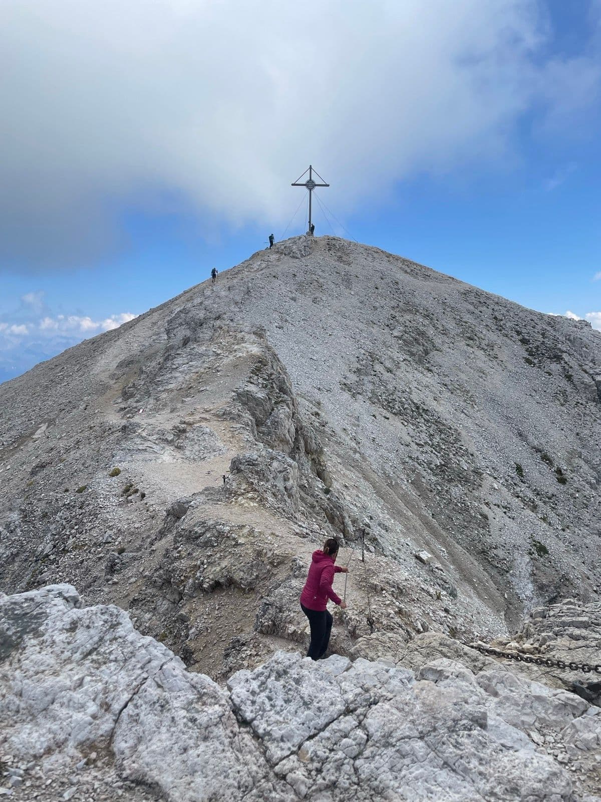Hiker climbing a rocky mountain ridge toward a summit cross in the Dolomites.