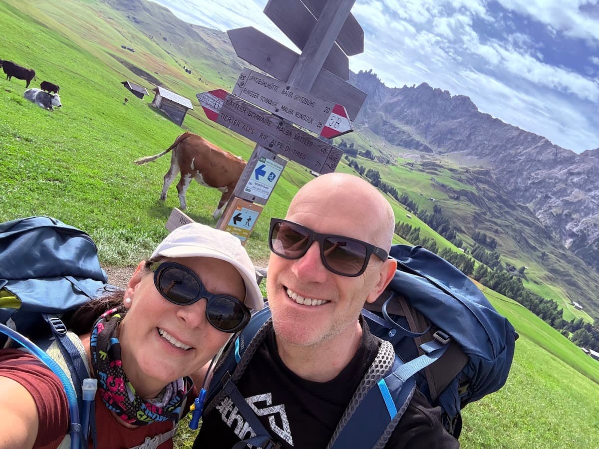 Hikers with backpacks taking a selfie near mountain trail signs and cows grazing in an alpine meadow