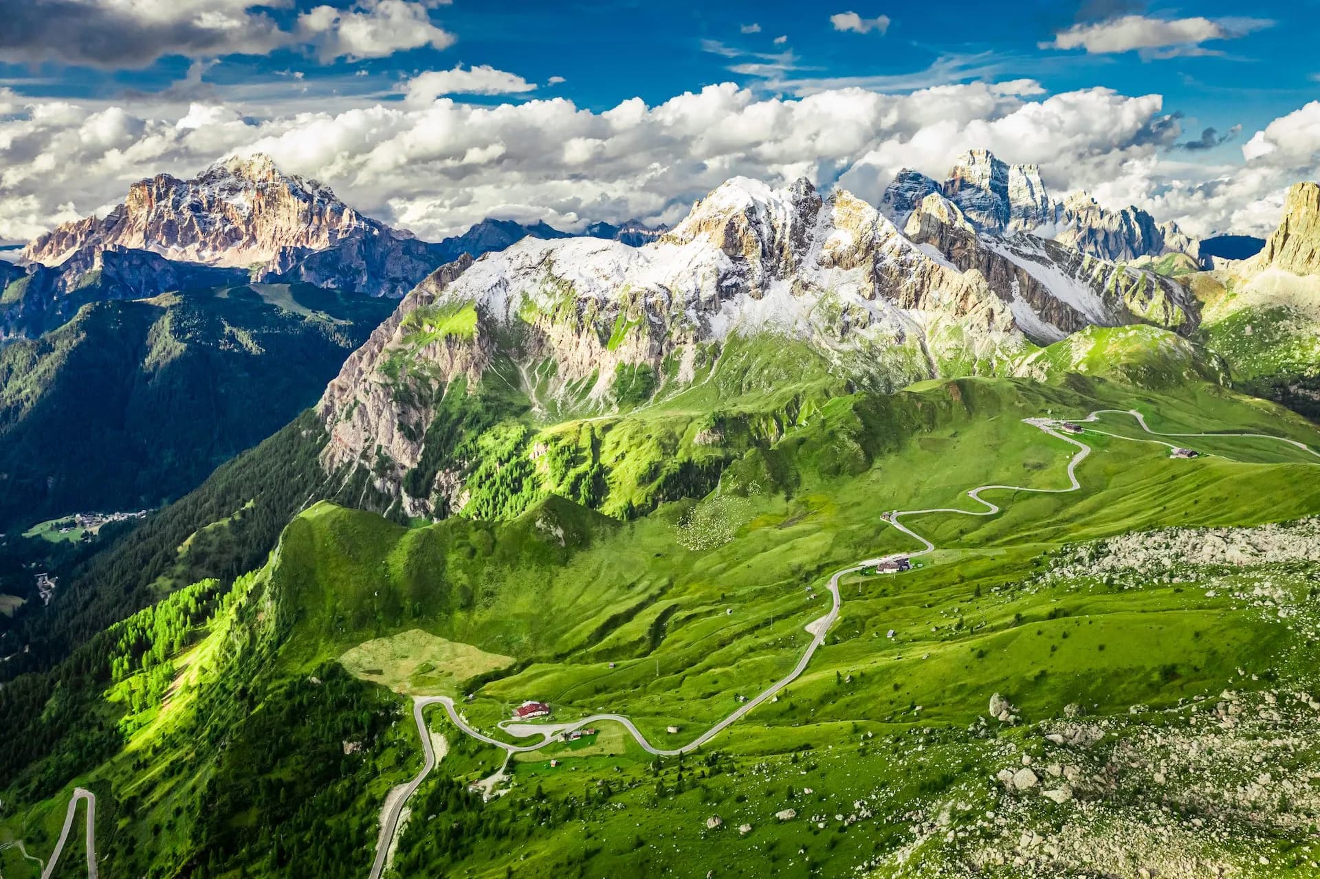 Winding mountain road through green slopes with snow-capped Dolomites peaks under a cloudy blue sky.