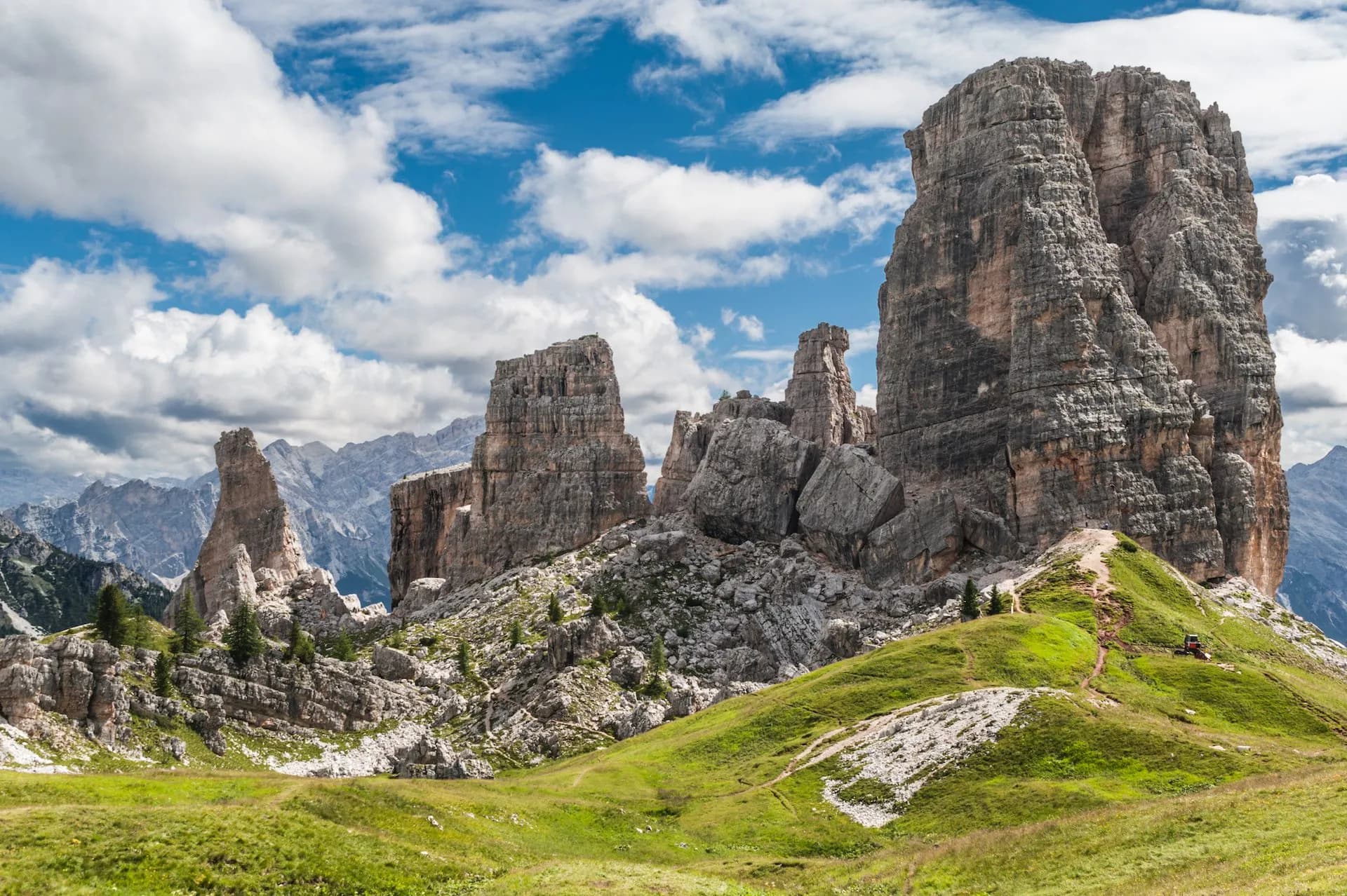 Majestic rock towers of Cinque Torri in the Dolomites with hiking trails and green meadows under a cloudy blue sky.