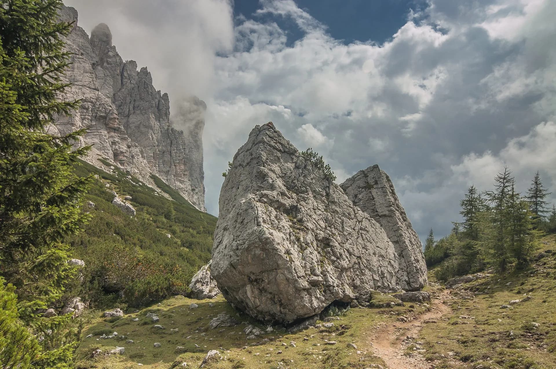 Hiking trail past large boulders toward towering, cloud-shrouded rock faces in the mountains.