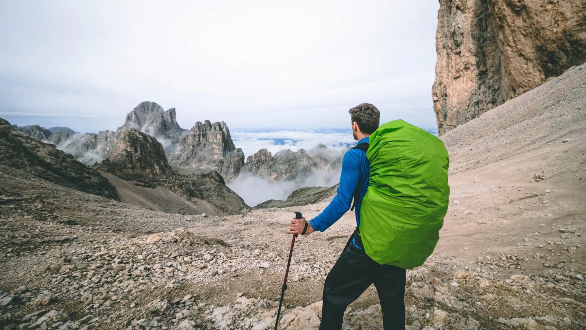 Hiker with green backpack and trekking pole overlooks rocky terrain and misty mountain peaks at Passo Antermoia.