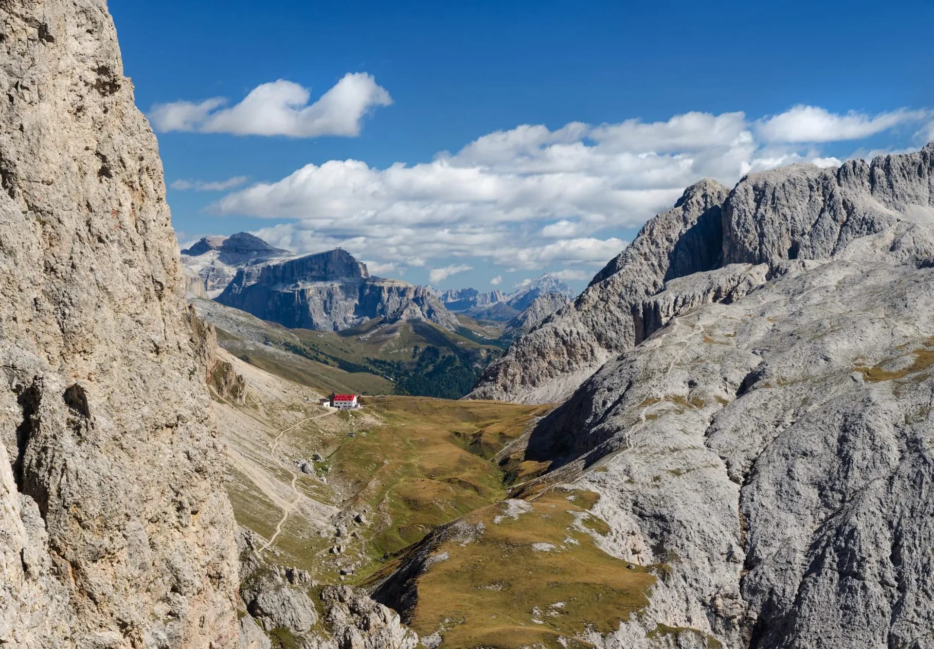 Mountain hut nestled in a valley surrounded by rugged, rocky peaks under a blue sky.