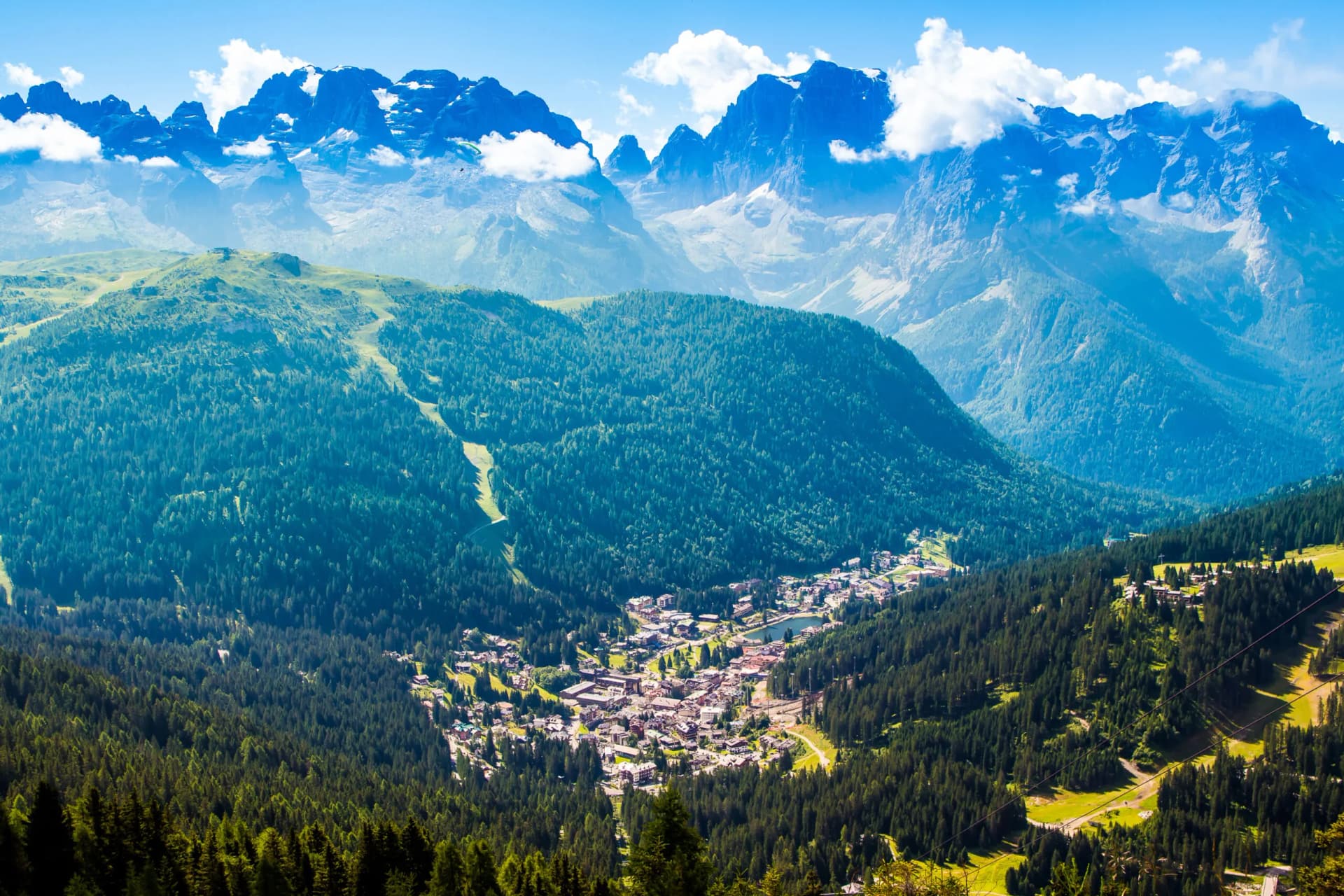 View of Madonna di Campiglio alpine village nestled in dense forest below jagged, snow-capped mountains.