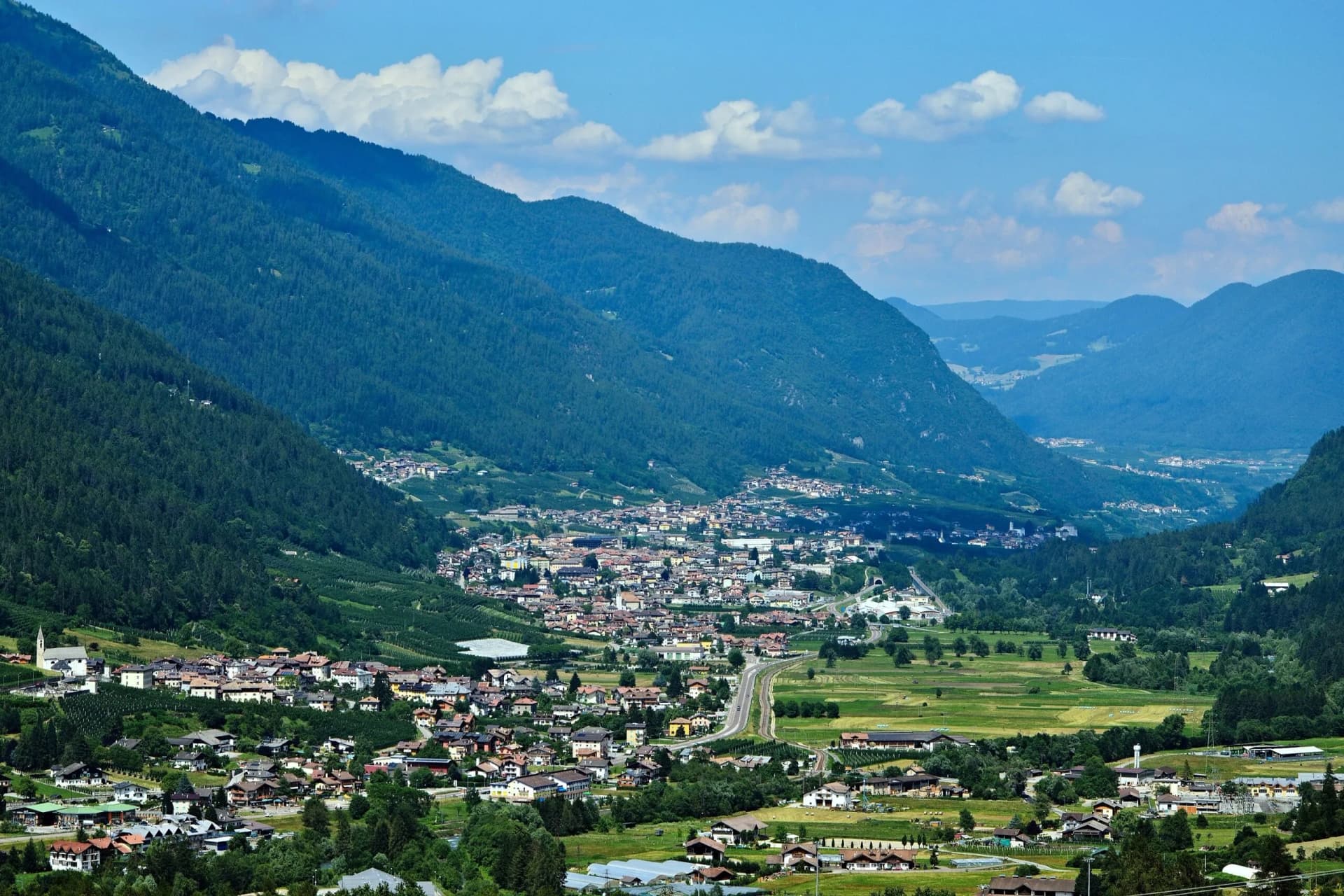 Alpine valley town nestled between steep, forested mountains under a blue, cloudy sky in Val di Sole.