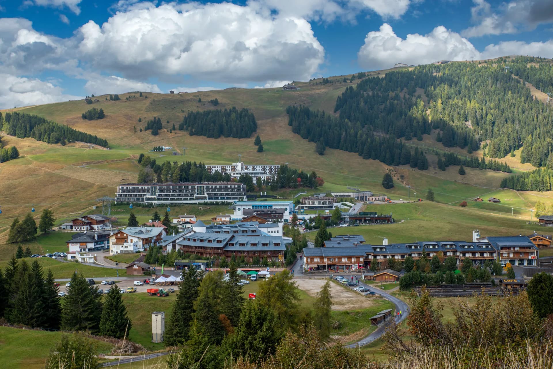 Alpine village with hotels nestled in green, rolling hills under a cloudy blue sky, Seiser Alm.